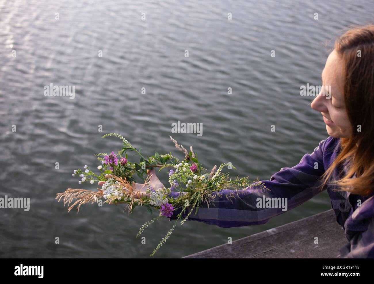 Die junge Frau bereitet sich darauf vor, einen Kranz mit Wildblumen ins Wasser zu werfen. Romantische Stimmung. Naturliebhaber. Traditionen für den Urlaub von Ivan Kupala. Enjo Stockfoto