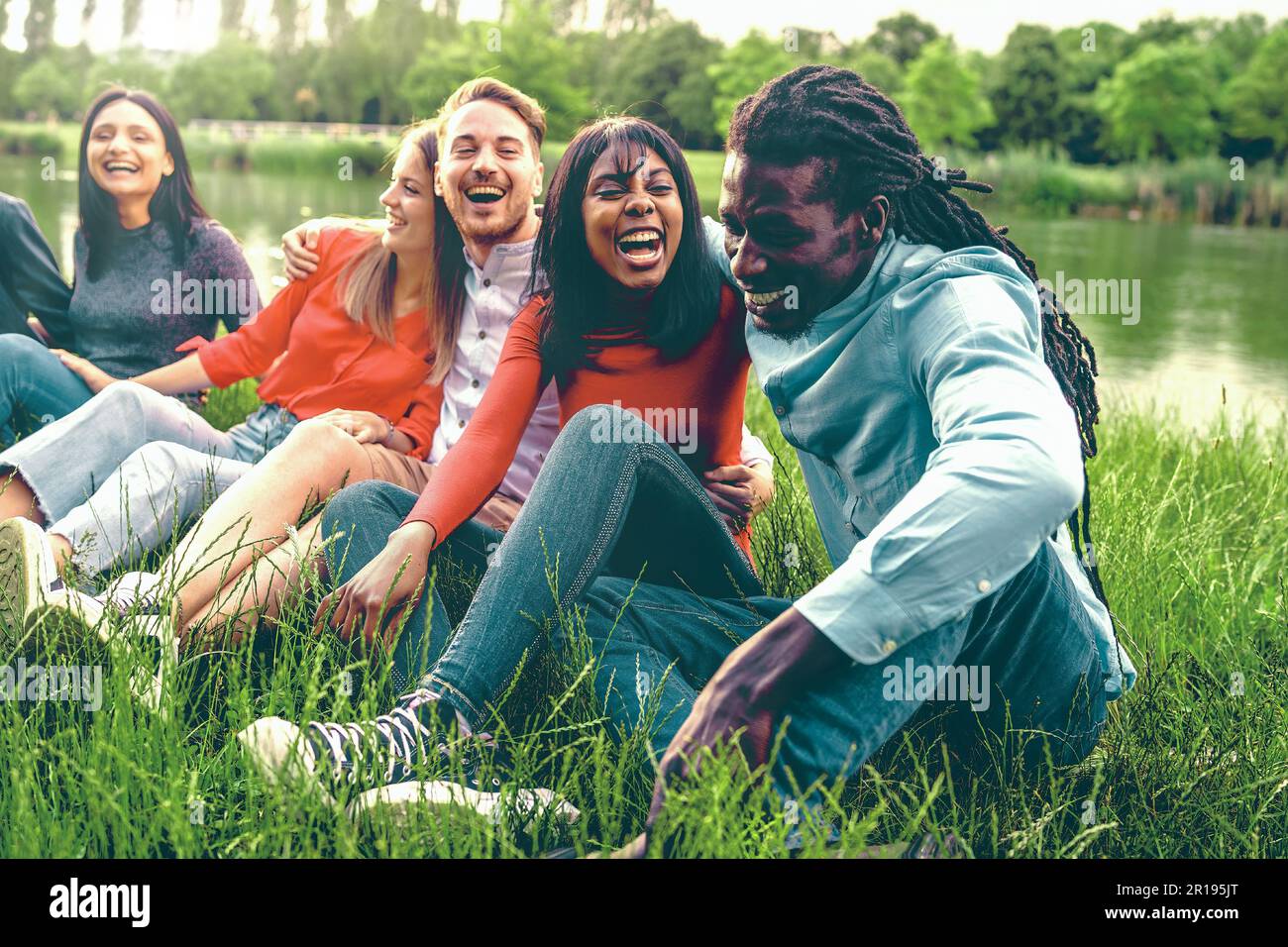 Eine Gruppe verschiedener junger Leute, die lachen und sich unterhalten, während sie auf dem Gras in einem Park in der Nähe eines Sees sitzen und Frühlings-/Sommerkleidung und emb tragen Stockfoto