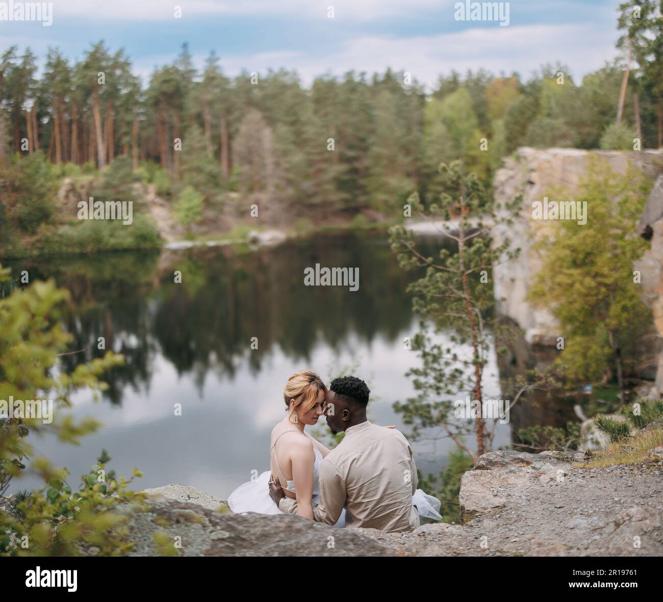 Glückliches gemischtrassiges Paar frisch Vermählte sitzen auf Felsen und umarmt sich vor dem wunderschönen Hintergrund des Sees, des Waldes und des Canyons. Konzept von Liebesbeziehungen und Stockfoto