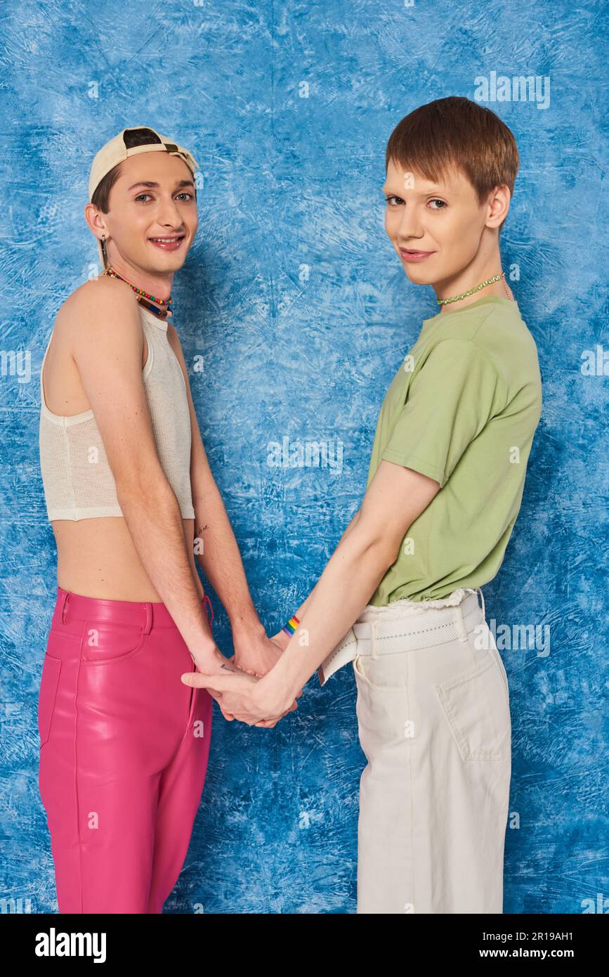 Lächelnde homosexuelle Freunde, die während der Homosexuellen-Monatsfeier Händchen halten und in die Kamera schauen, auf strukturiertem und meliertem blauem Hintergrund Stockfoto
