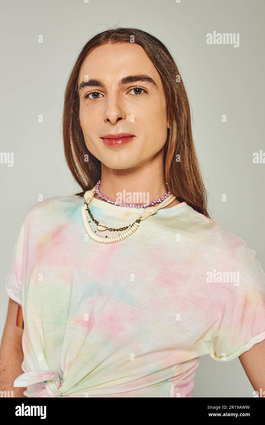 Porträt eines jungen und langhaarigen homosexuellen Mannes in einem T-Shirt mit Krawatte und Blick auf die Kamera isoliert auf grauem, Happy Pride Community Month Feier Konzert Stockfoto