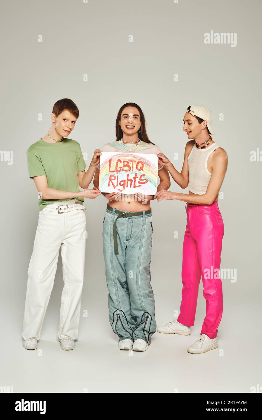 Lächelnde und junge, nicht binäre Freunde halten ein Plakat mit lgbtq-Schriftzug und stehen zusammen, während sie den Pride Community Month auf Grau feiern Stockfoto
