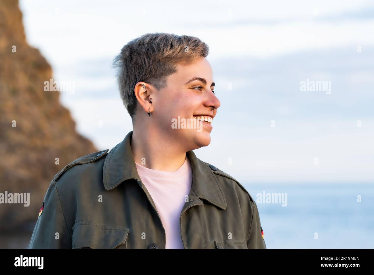 Porträt einer nicht-binären Person mit kurzen Haaren, leicht kurvig, am Meer lächelnd. Sie tragen ein rosa Hemd und eine grüne Militärjacke Stockfoto