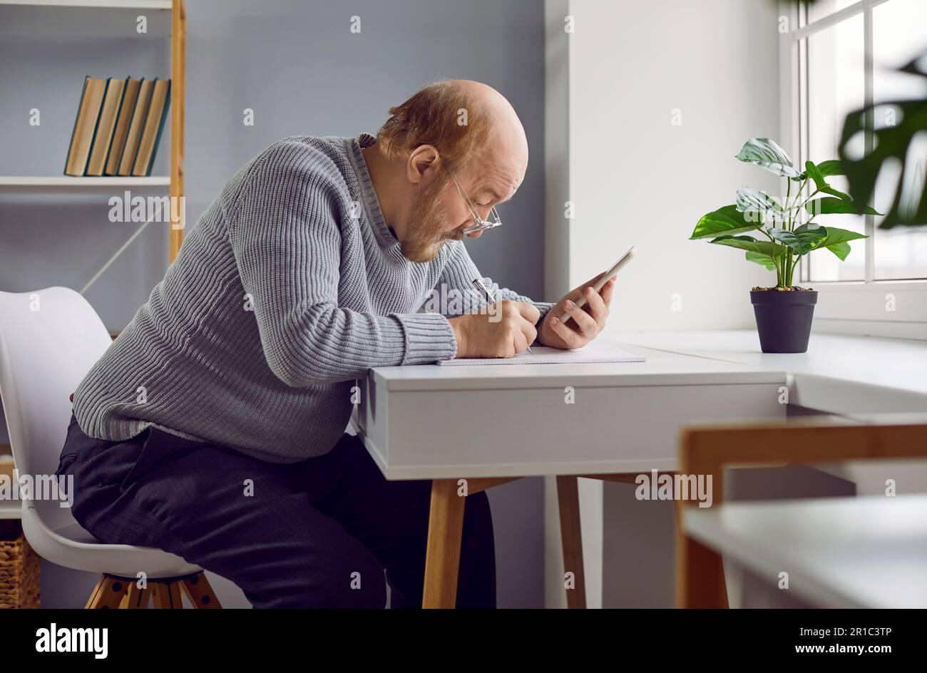 Ein älterer Mann, der zu Hause am Tisch sitzt, sein Handy ansieht und sich Notizen macht Stockfoto