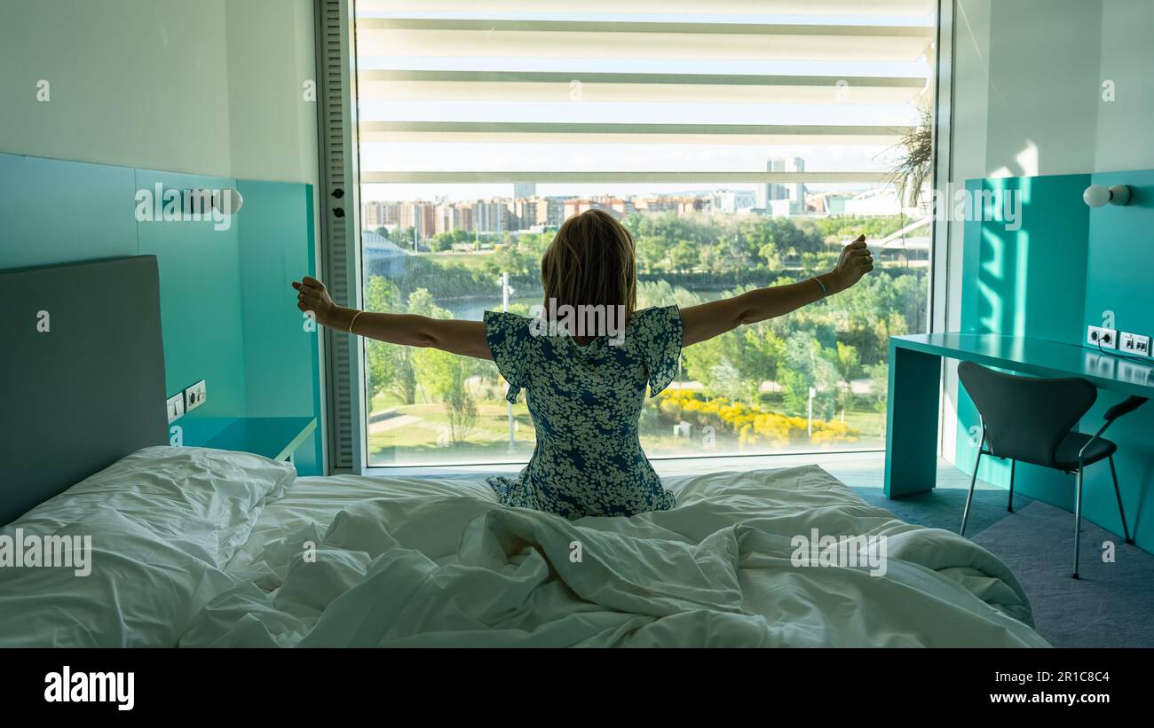 Frau, die sich streckt und auf dem Bett sitzt, nachdem sie vor einem großen Fenster mit Aussicht aufgewacht ist. Stockfoto