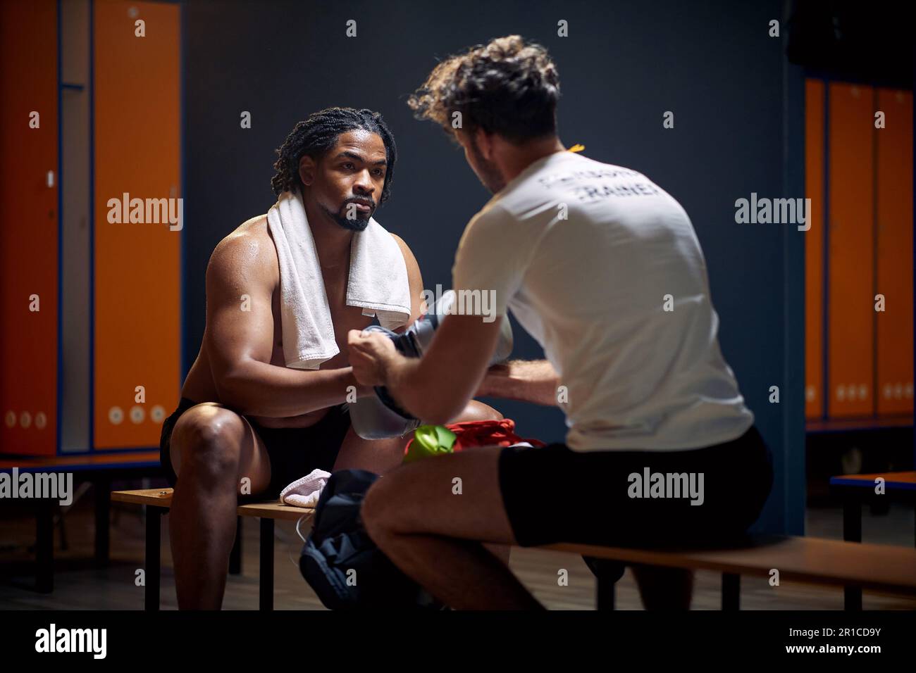Starker junger Boxspieler nach dem Workout mit dem Trainer, der ihm dabei hilft, die Handschuhe auszuziehen, und der sich im Gespräch von Angesicht zu Angesicht in der Garderobe aufhält. Sport, Lifestyle Stockfoto