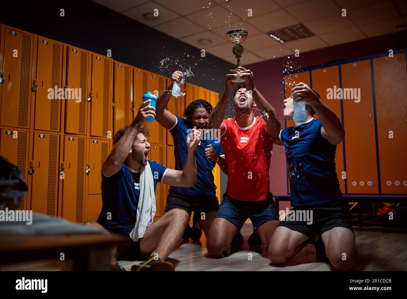 Eine Gruppe von Basketballspielern hält die goldene Trophäe hoch und feiert den Erfolg in der Garderobe, schreit und spritzt Wasser. Erster Platz gewonnen. Stockfoto