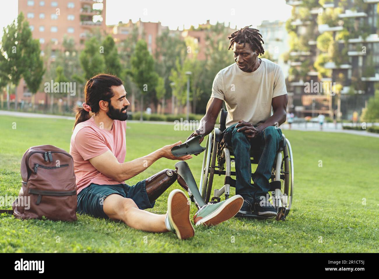 Ein hispanischer Mann mit Bart, Pferdeschwanz und Beinprothese sitzt auf dem Gras in einem öffentlichen Park und zeigt seinem afrikanischen Freund im Rollstuhl ein Stück von einem Stockfoto