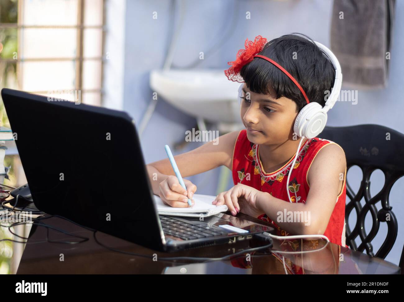 Ein süßes indisches Mädchen in rotem Kleid, das zu Hause mit Laptop und Kopfhörer lernt Stockfoto
