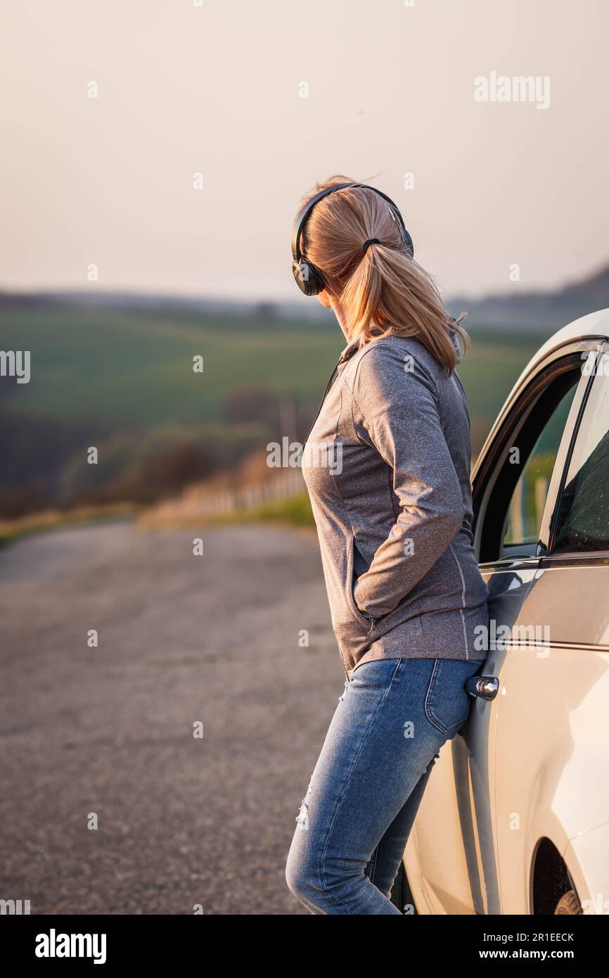 Frau mit Kopfhörern, die sich auf dem Auto anlehnte und sich während einer Autofahrt ausruhte. Weibliche Person, die Alleinreisende genießt Stockfoto