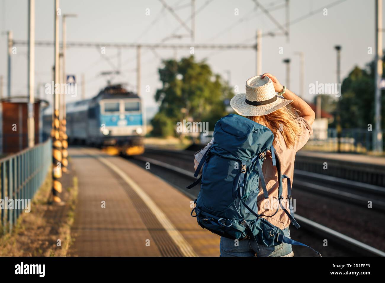 Weibliche Rucksacktouristin, die allein mit dem Zug reist. Frau mit Rucksack und Strohhut, die am Bahnhofsplatz steht und auf den ankommenden Zug schaut Stockfoto