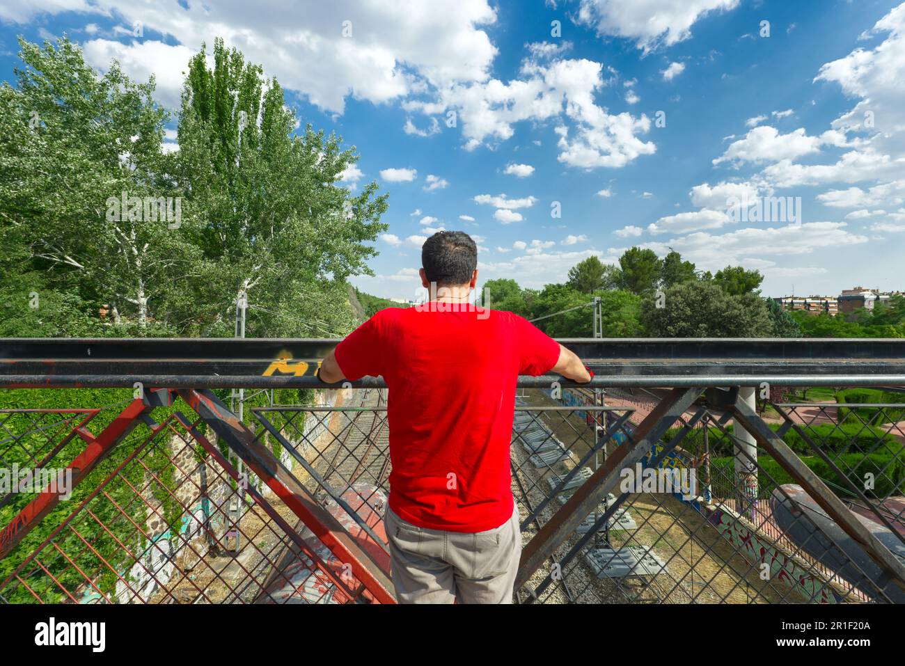 Ich habe mich selbst dargestellt, als ich die Gleise von einer Brücke mit einem hohen Stahlgeländer an einem Tag des hellen Himmels mit ein paar Wolken betrachtete Stockfoto