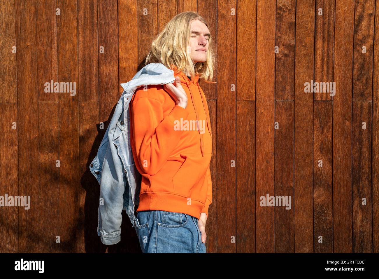 Der entspannte, blonde Mann in der Jacke über der Schulter mit geschlossenen Augen steht in der Sonne an der Holzwand Stockfoto