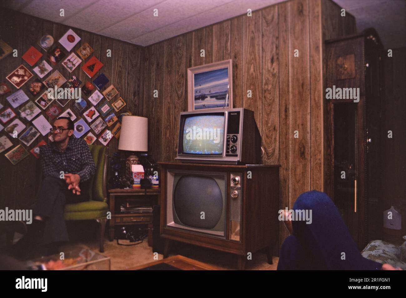 Typisches Wohnzimmer in einem amerikanischen Heim zu Weihnachten, Weihnachtskarten hängen an einer holzgetäfelten Wand und mit einem kaputten Konsolenfernseher und einem kleineren Fernseher darüber. 1982 Stockfoto
