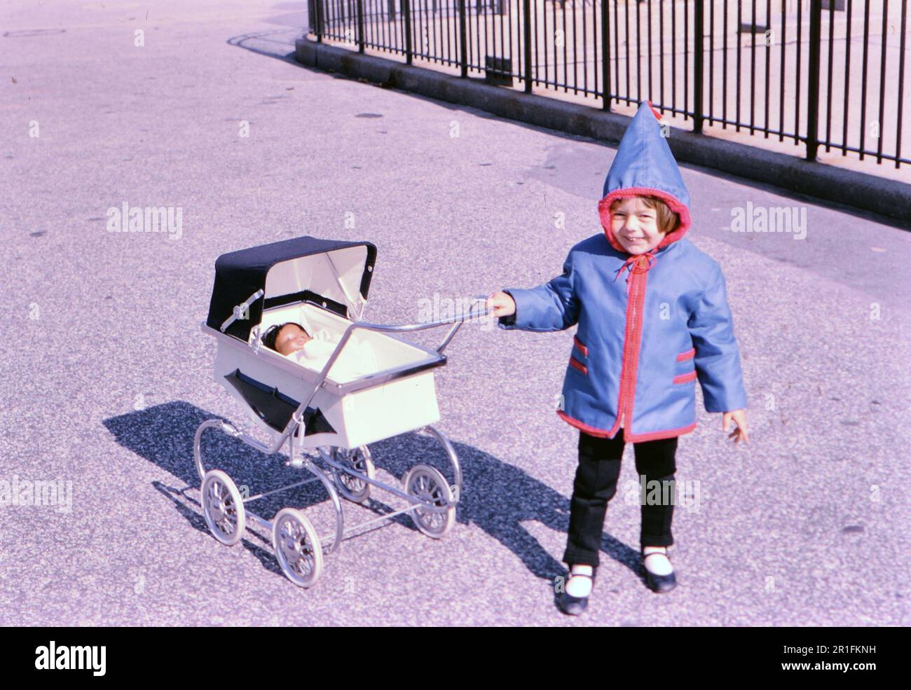 Ein kleines Mädchen an einem kalten Tag, das einen Mantel trägt und einen Kinderwagen schiebt, mit ihrer Spielzeugpuppe in ca. 1960er Stockfoto