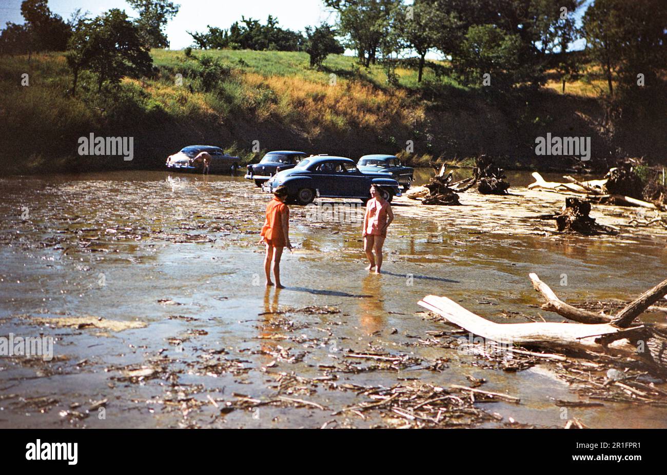 Zwei Mädchen, die in einem Fluss spazieren (wahrscheinlich im Hügelgebiet von Texas), Autos, die auf einem flachen Felsbett in Niedrigwasser geparkt sind, ca. 1954 Stockfoto