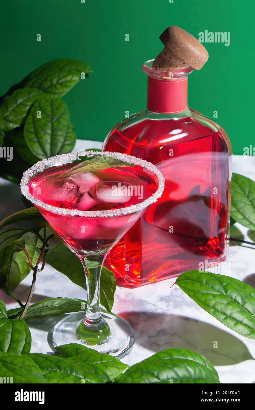 Roter Cocktail mit Eis in einem margarita-Glas mit einem Zuckerrand,  daneben ist roter Alkohol in einer Flasche mit einem Holzkorken. Umgeben  von grünen Blättern von Stockfotografie - Alamy