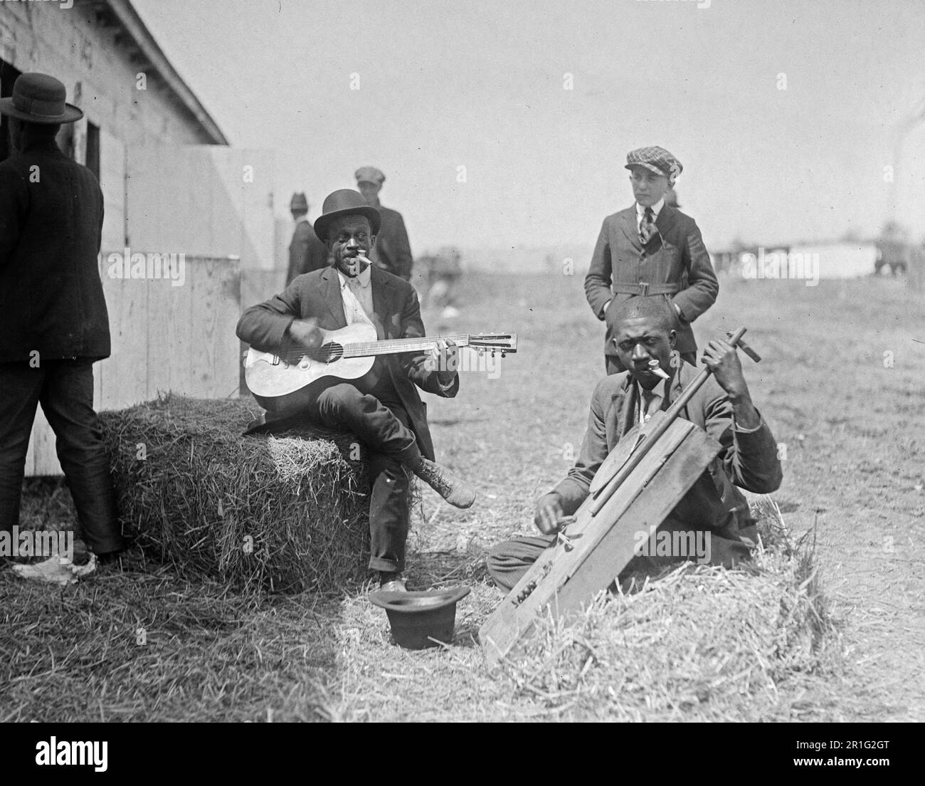 Archivfoto: Das Foto zeigt zwei Männer, wahrscheinlich Afroamerikaner, die auf Heuballen sitzen und vor einer Scheune oder einem Stall Instrumente spielen. Ein Mann spielt Gitarre, der andere spielt ein Instrument, das einem Cello ähnelt; beide Männer spielen gleichzeitig Kazoos Ca. 1918-1920 Stockfoto