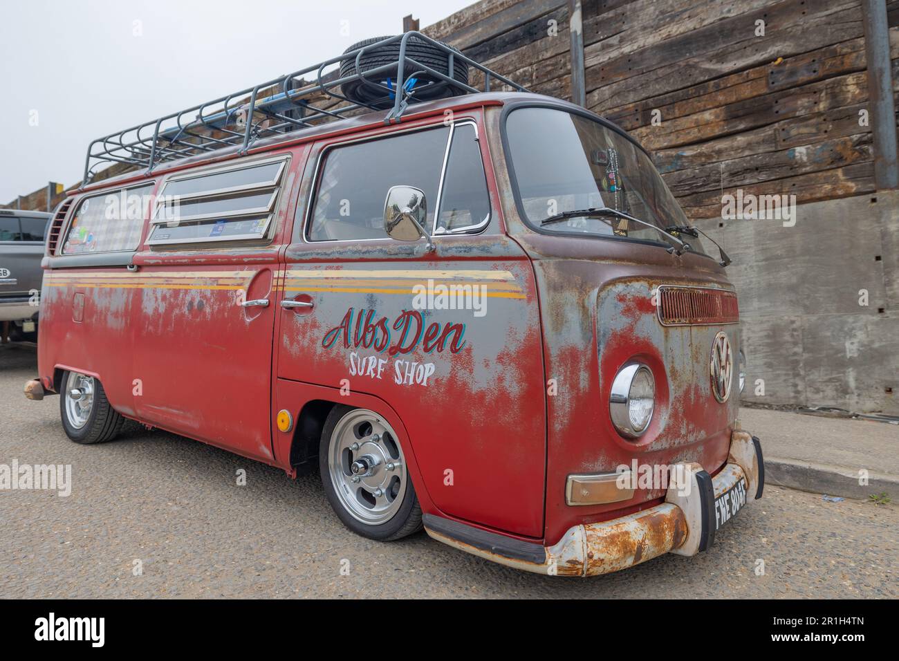 Rayleigh, Großbritannien. Mai 2023. Ein verwitterter roter Volkswagen Typ 2 Van mit Surf Shop Branding („AltsDen Surf Shop“) parkt neben einer holzverkleideten Mauer in Rayleigh, Essex. Der Van verfügt über eine abgesenkte Federung, verchromte Räder und einen Dachträger mit Spiralkabel, der an nostalgische Surfkultur und ein individuelles Auto-Styling erinnert. Farm Fresh Garage Truck und amerikanische Fahrzeuge werden gezeigt. Penelope Barritt/Alamy Live News Stockfoto