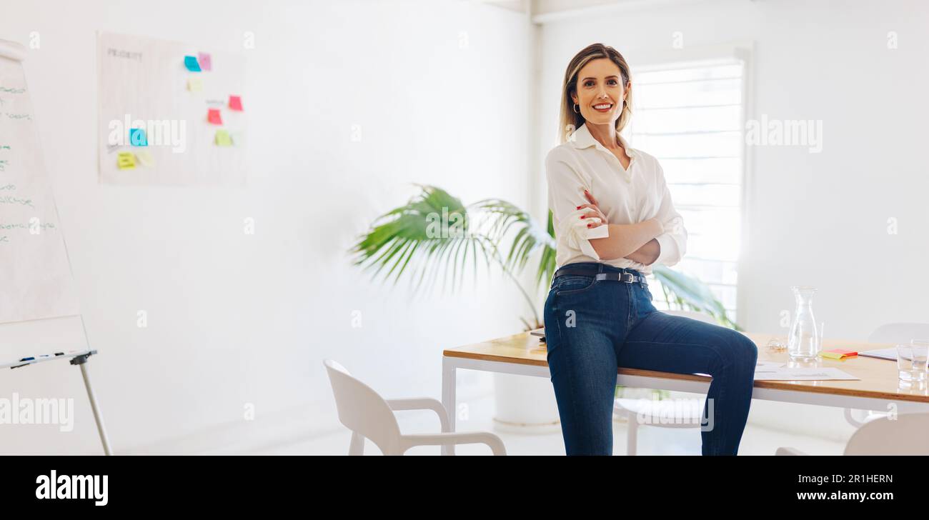 Kreative Geschäftsfrau, die vor der Kamera lächelt, während sie auf einem Konferenztisch in einem Sitzungssaal sitzt. Fröhliche Unternehmerin, die in einem modernen Werk arbeitet Stockfoto