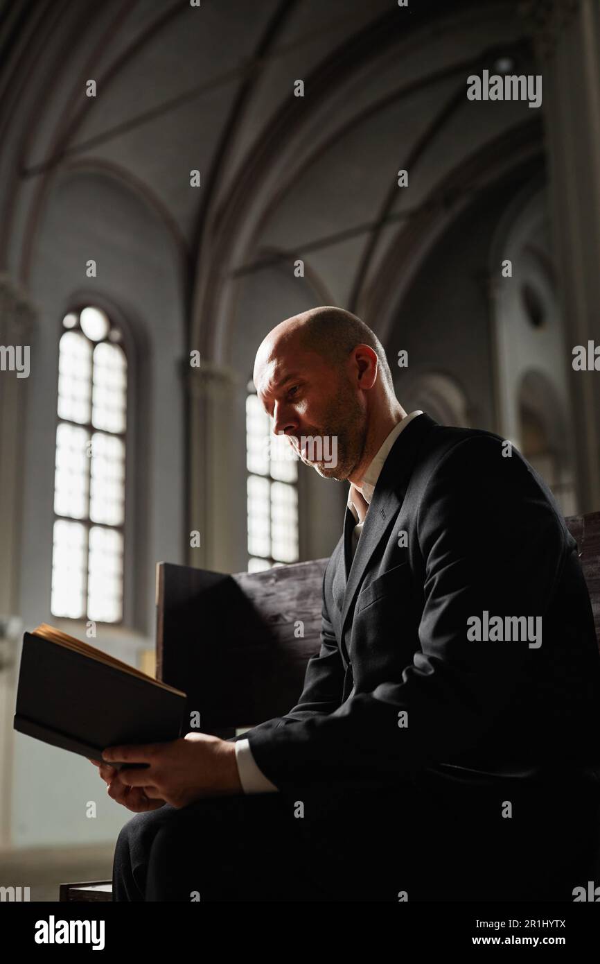 Ein erwachsener Mann im schwarzen Anzug, der auf einer Bank sitzt und Gebete aus der Bibel in der Kirche liest Stockfoto