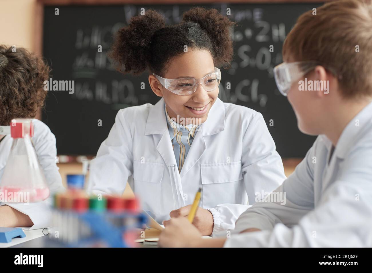 Portrait eines lächelnden schwarzen Schulmädchens, das im Wissenschaftsunterricht einen Laborkittel trägt und mit einer Freundin spricht. Stockfoto