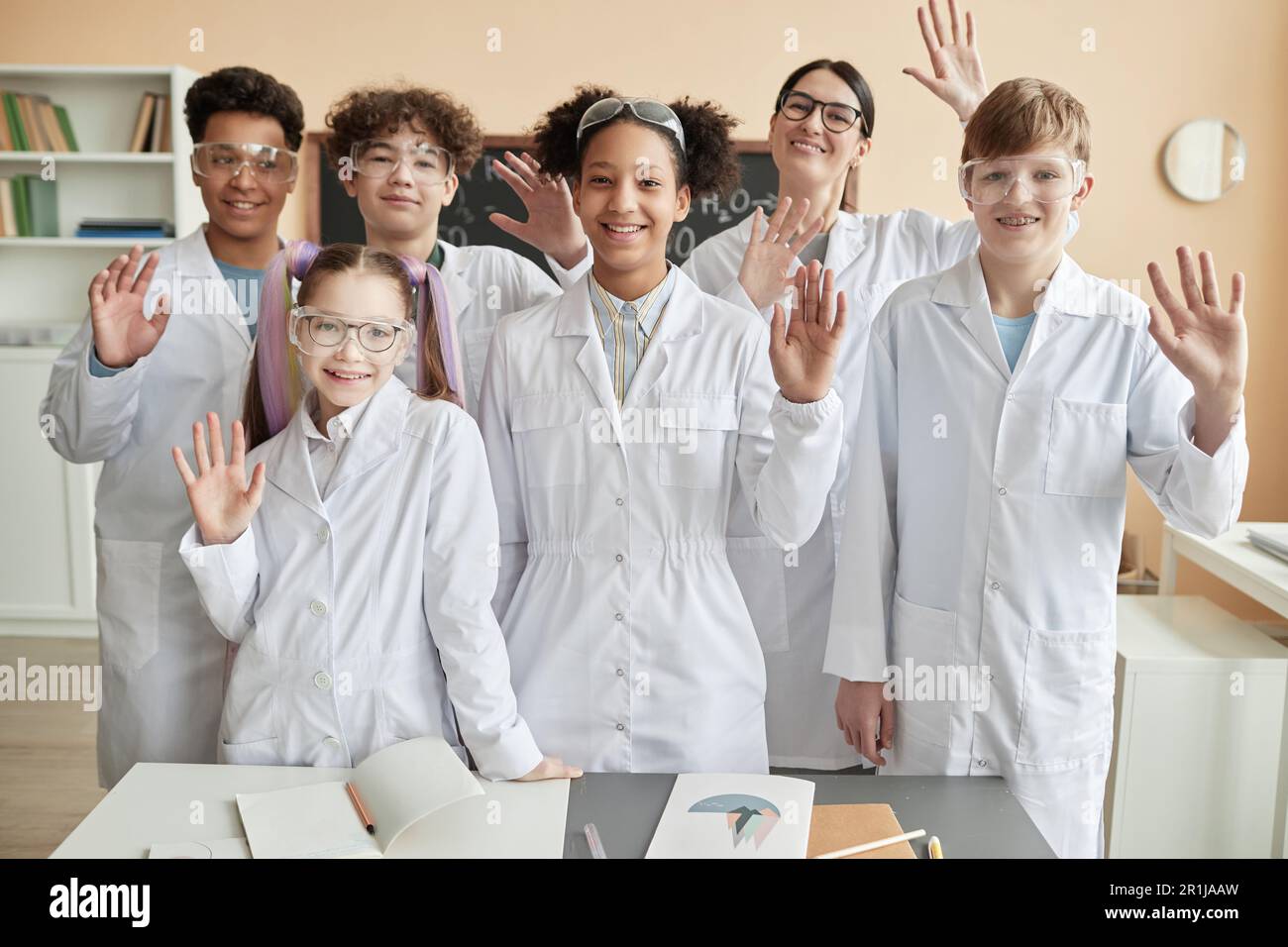 Eine vielfältige Gruppe glücklicher Teenager, die in der Schule Labormäntel tragen und vor der Kamera winken, zusammen stehen Stockfoto