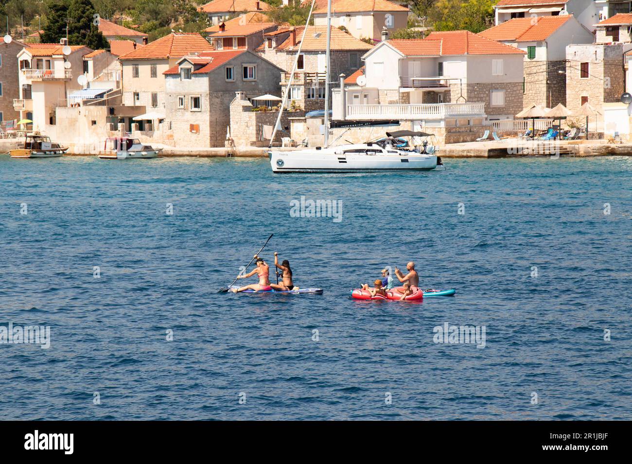 Prvic Luka, Kroatien - 22. Juli 2022: Leute paddeln, während sie auf Paddleboards und Steinhäusern am Dock dahinter sitzen Stockfoto