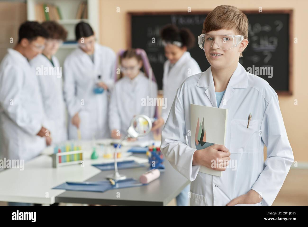 Porträt eines Teenager-Schülers, der einen Laborkittel im Wissenschaftsunterricht trägt und mit einer Gruppe von Kindern im Hintergrund in die Kamera schaut, Kopierraum Stockfoto