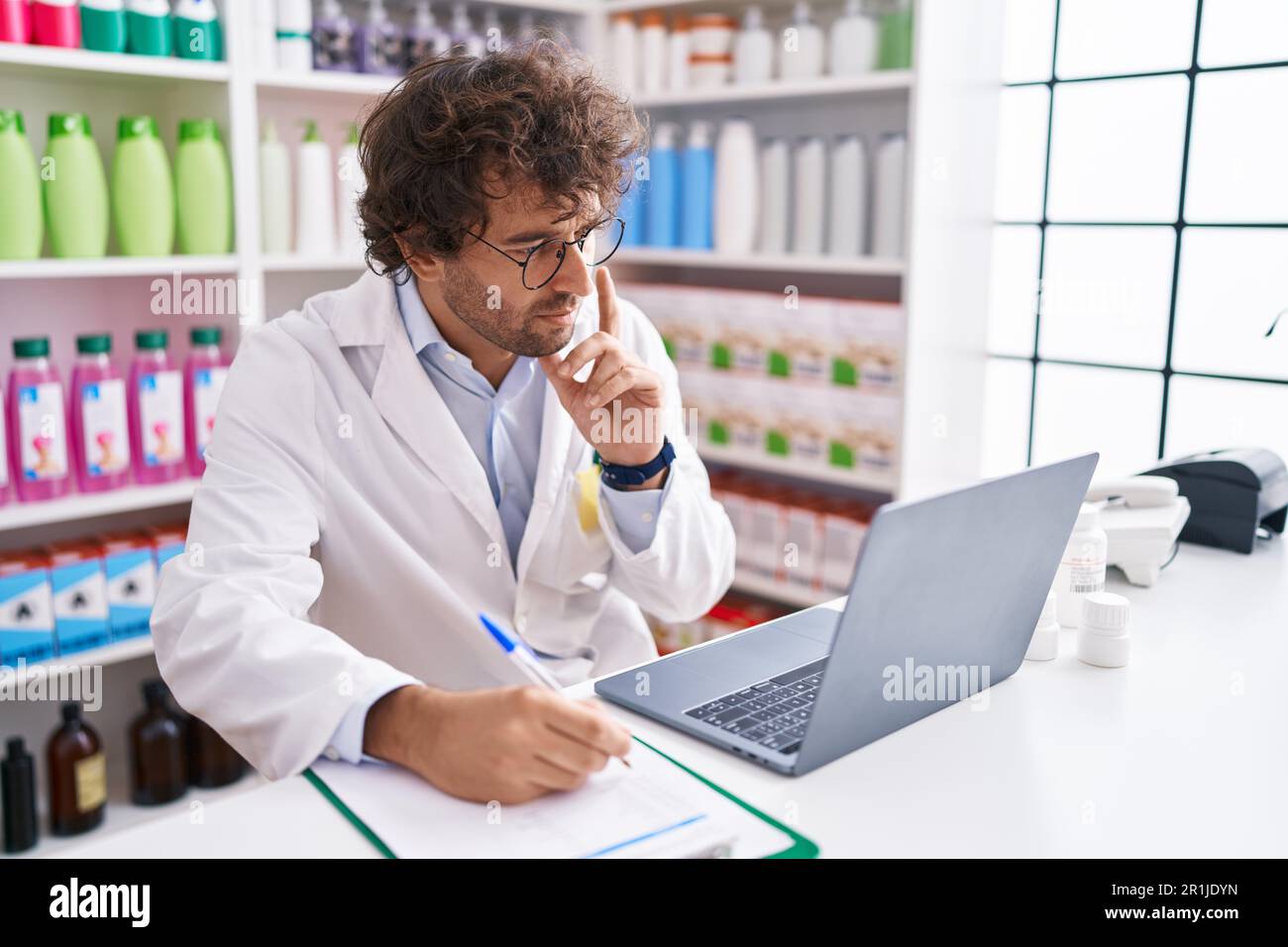Junger hispanischer Apotheker, der mit dem Laptop in der Apotheke auf Dokumente schreibt Stockfoto