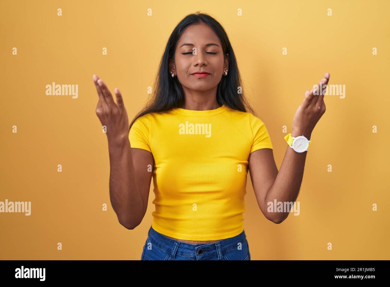 Junge indianerin, die auf gelbem Hintergrund steht, entspannt und mit geschlossenen Augen lächelt und Meditationsgeste mit den Fingern macht. Yoga-Konzept. Stockfoto