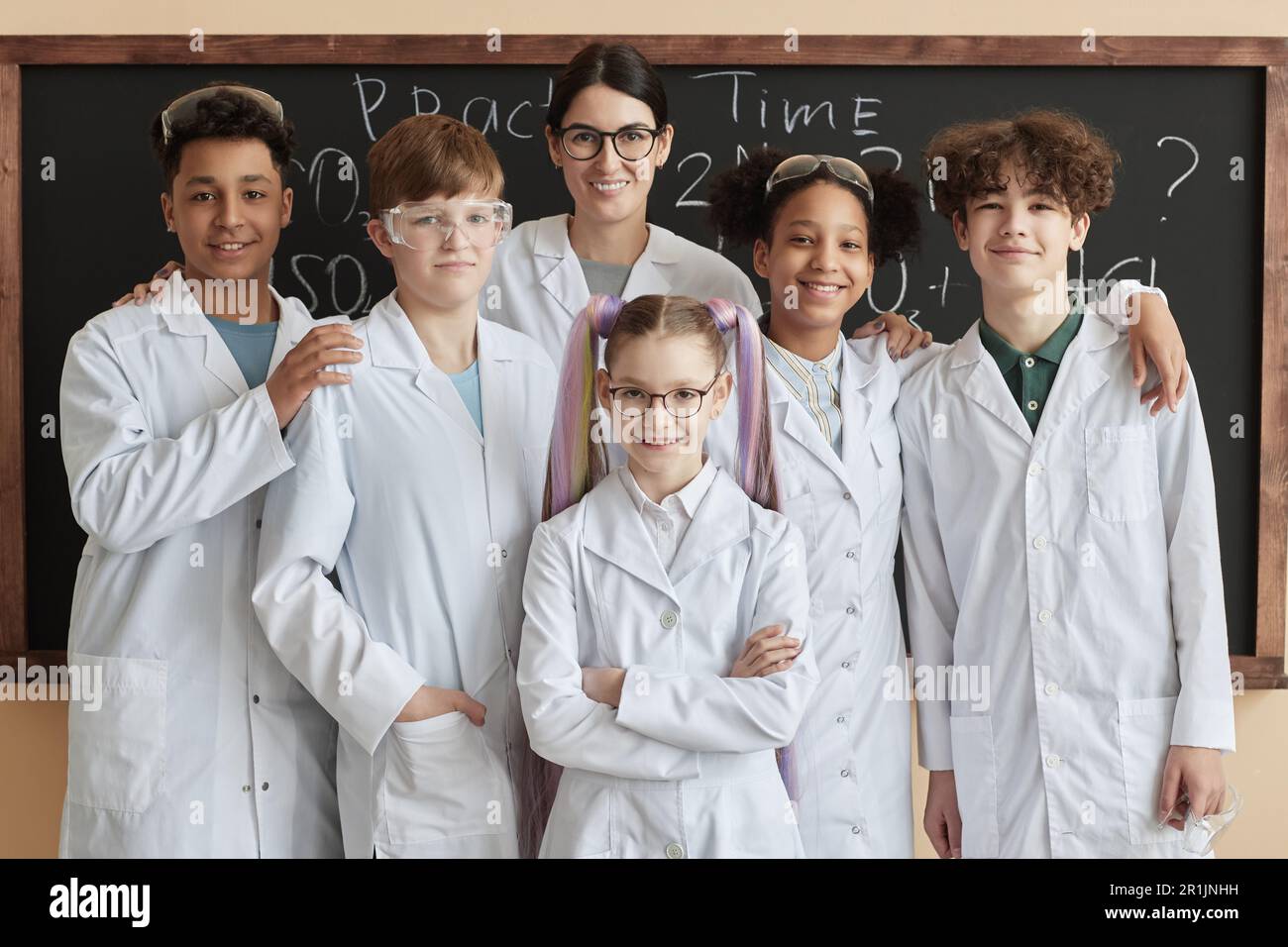 Eine vielfältige Gruppe von Kindern, die Labormäntel tragen und mit einem Lehrer in der Wissenschaftsklasse posieren und vor der Kamera lächeln Stockfoto