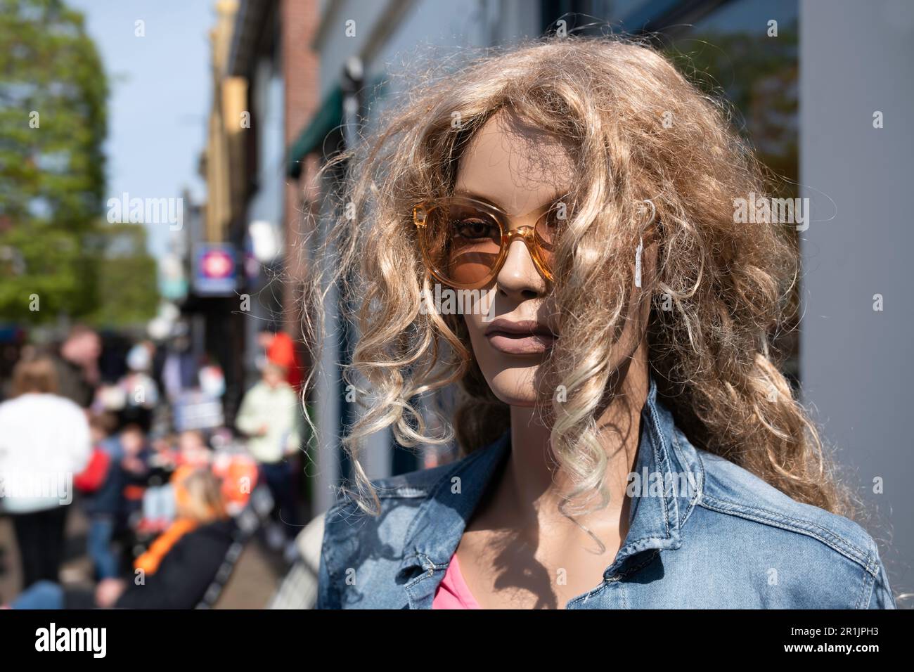 Eine weibliche Plastikpuppe mit lockiger Perücke und Sonnenbrille steht vor der Fassade eines Modegeschäfts in einer Einkaufsstraße Stockfoto