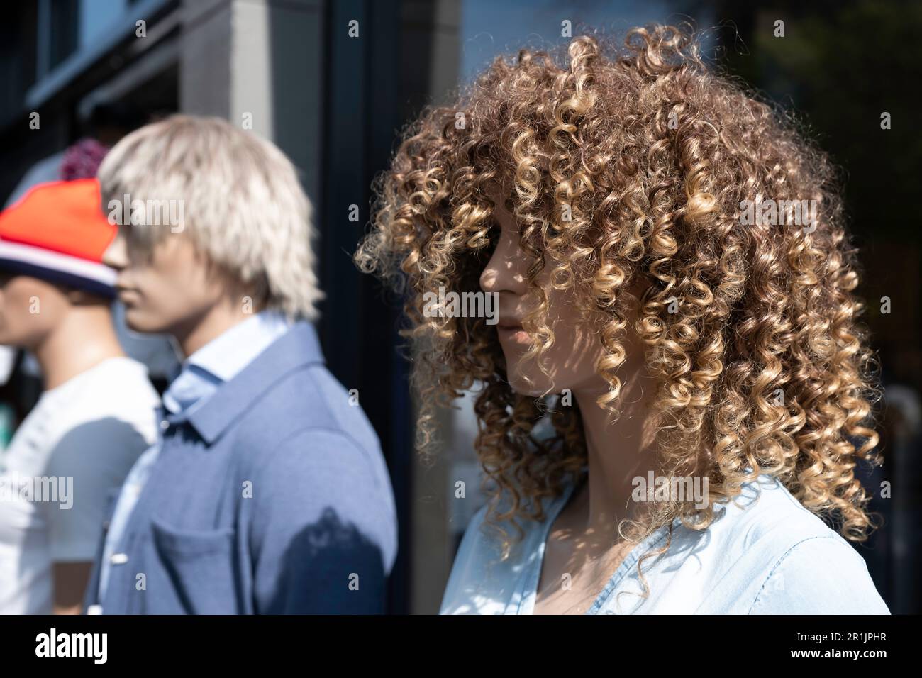 Drei Schaufensterpuppen aus Kunststoff stehen nebeneinander vor der Fassade eines Modegeschäfts. Konzentrier dich auf den Richtigen mit einer lockigen Perücke Stockfoto