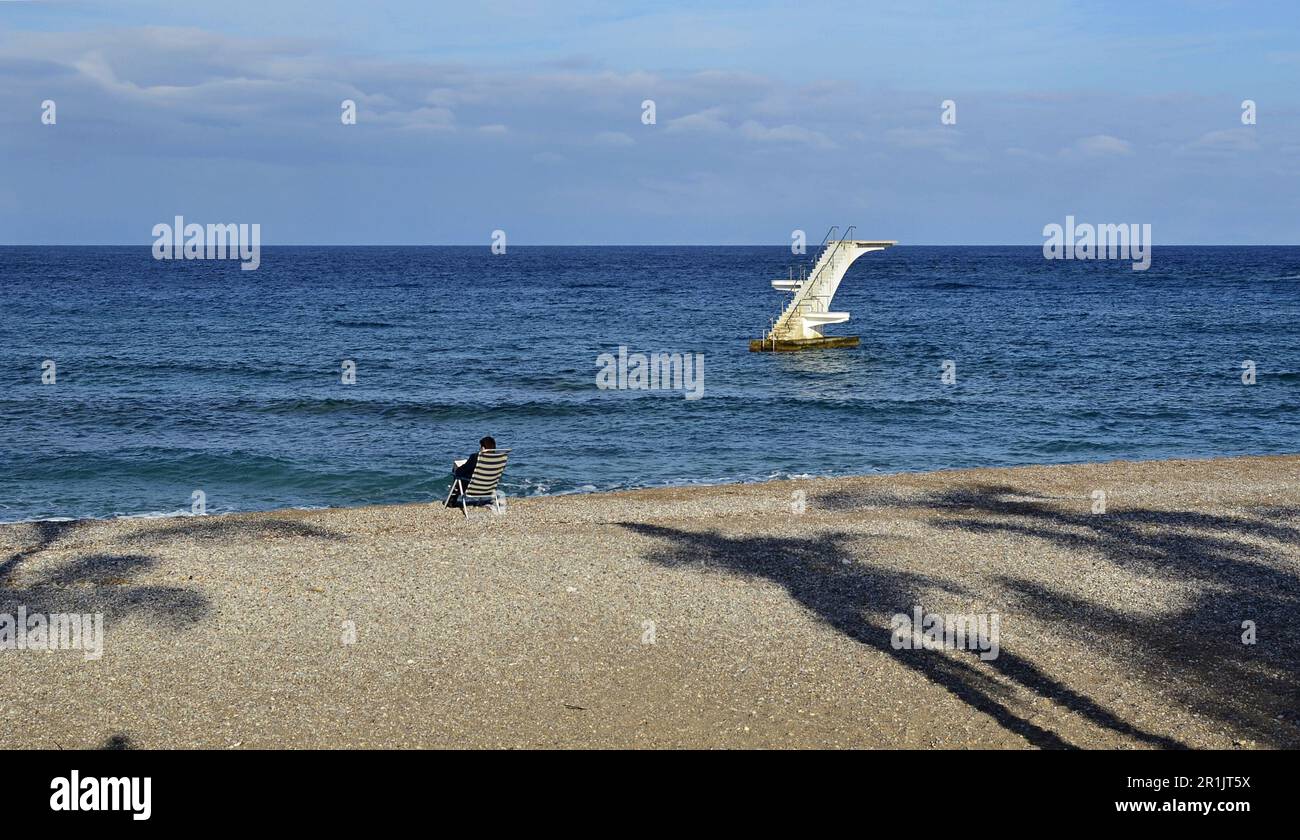 Seeküste mit Kieselstrand. Verlassene Winterküste. Ein Tauchturm ist im Meer sichtbar. Ein Mann in einem Liegestuhl sitzt am Ufer Stockfoto