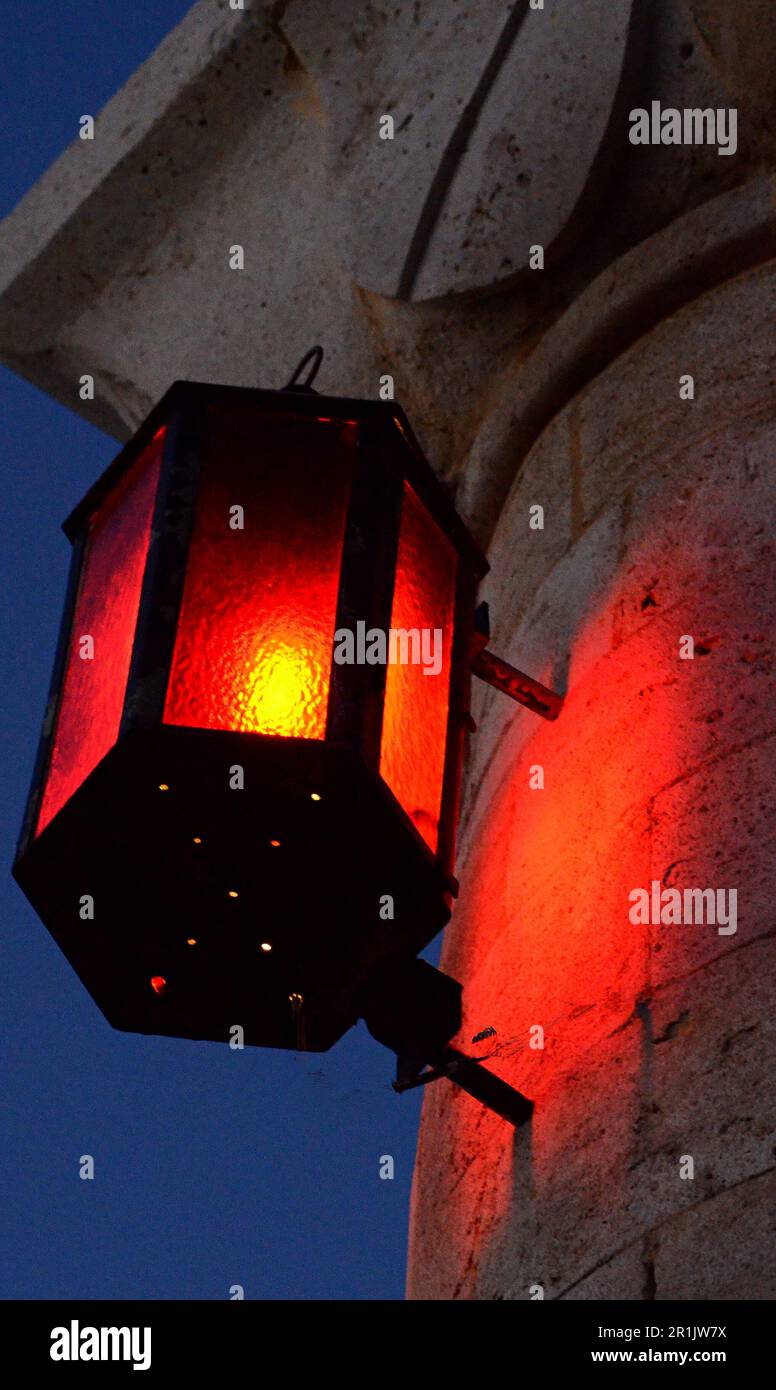 Eine rote Laterne in einem Metallrahmen wird am Abend auf einer Steinsäule am Eingang zum Hafen der Insel beleuchtet. Die Laterne ist aus nächster Nähe sichtbar. Stockfoto