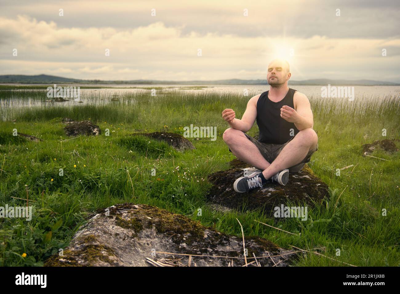 Wunderschöne Naturlandschaft mit Mann in ärmellosem Hemd und Camo-Shorts beim Meditieren auf dem Felsen am Corrib Lake in Galway, Irland Stockfoto