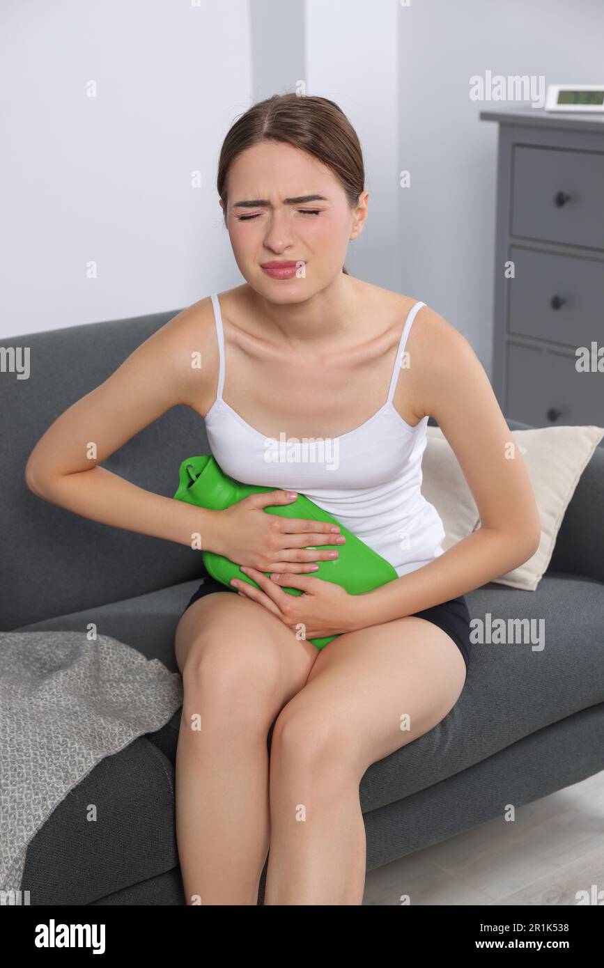 Junge Frau, die zu Hause eine Wärmflasche benutzt, um Zystitisschmerzen auf dem Sofa zu lindern Stockfoto