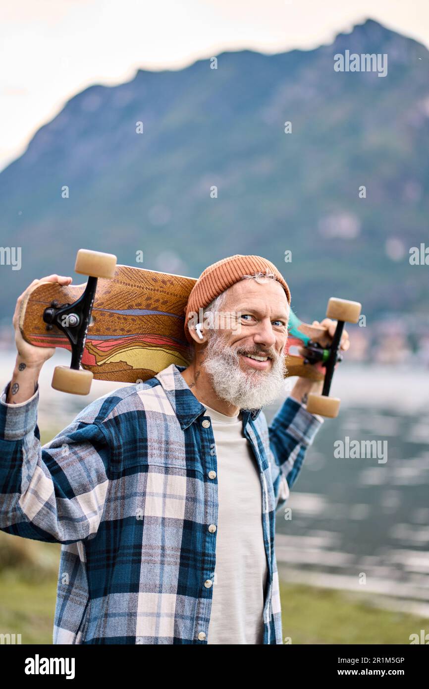 Ein älterer, glücklicher Hipster-Skater steht im Naturpark und hält Schlittschuh. Stockfoto