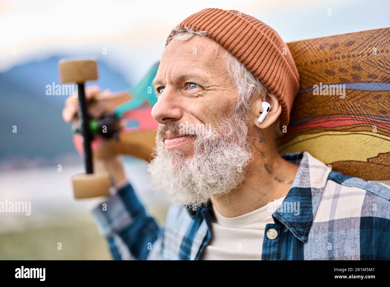 Ein älterer Hipster, der im Naturpark steht und Skateboard hält und Musik hört. Stockfoto