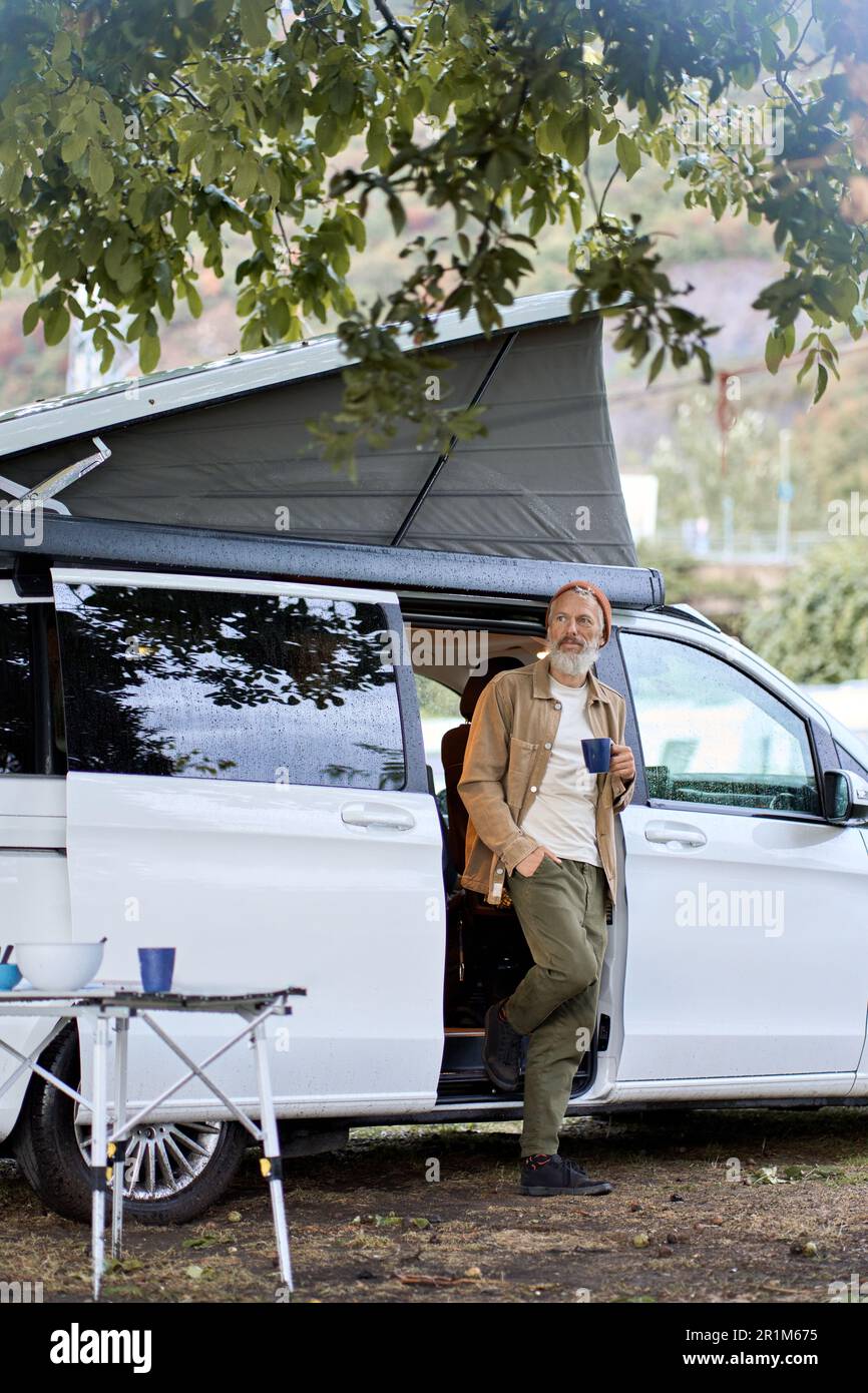 Aktiver älterer Hipstermann, der in der Nähe eines Wohnwagens auf einer Urlaubsreise steht. Stockfoto