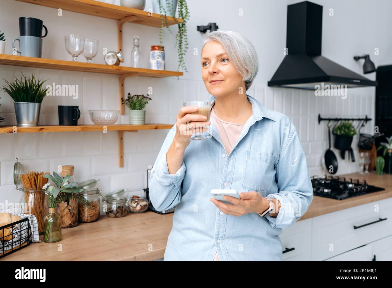Wunderschöne weiße grauhaarige Dame mittleren Alters, steht zu Hause in der Küche, hält ihr Smartphone und ein Glas Kaffee, chattet in sozialen Netzwerken, liest Nachrichten, sieht weg, träumt, lächelt Stockfoto