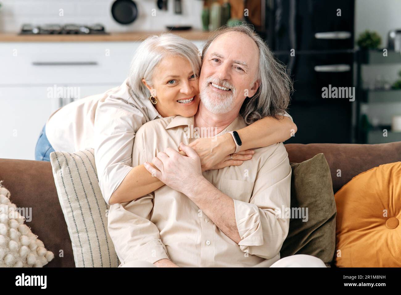 Positiv glückliches weißes, reifes Ehepaar, grauhaarige Ehepartner verbringen Zeit zusammen in einem Wohnzimmer zu Hause, Frau umarmt ihren Mann auf dem Sofa, sie schauen in die Kamera, lächeln Stockfoto