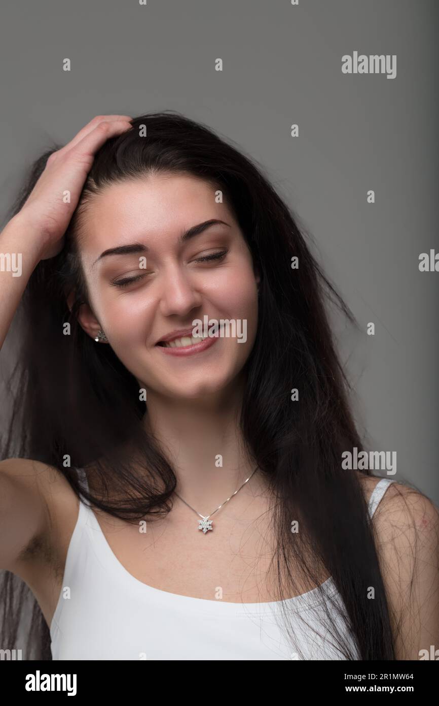 Lächelnde Frau, weißes Tanktop, langes dunkles Haar, sorglose Jugend, Körperpflege, Make-up, Schmuckkette, fesselnde Pose Stockfoto