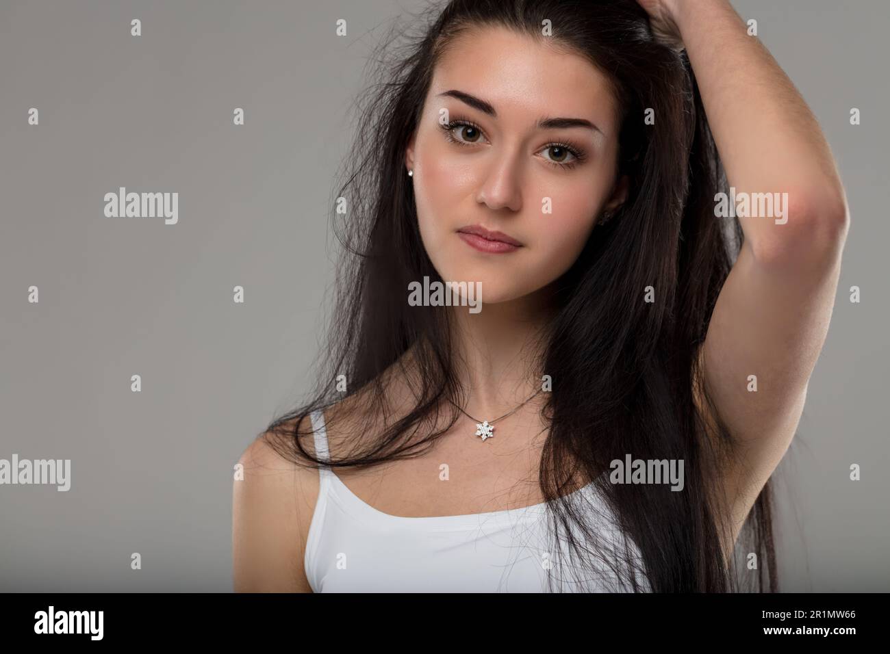 Junge Frau in weißem Tanktop, Hand in schwarzem Haar, lächelnd, sorgenfrei, gut gepflegt, Make-up, Halskette mit Juwel, intensiver Blick Stockfoto