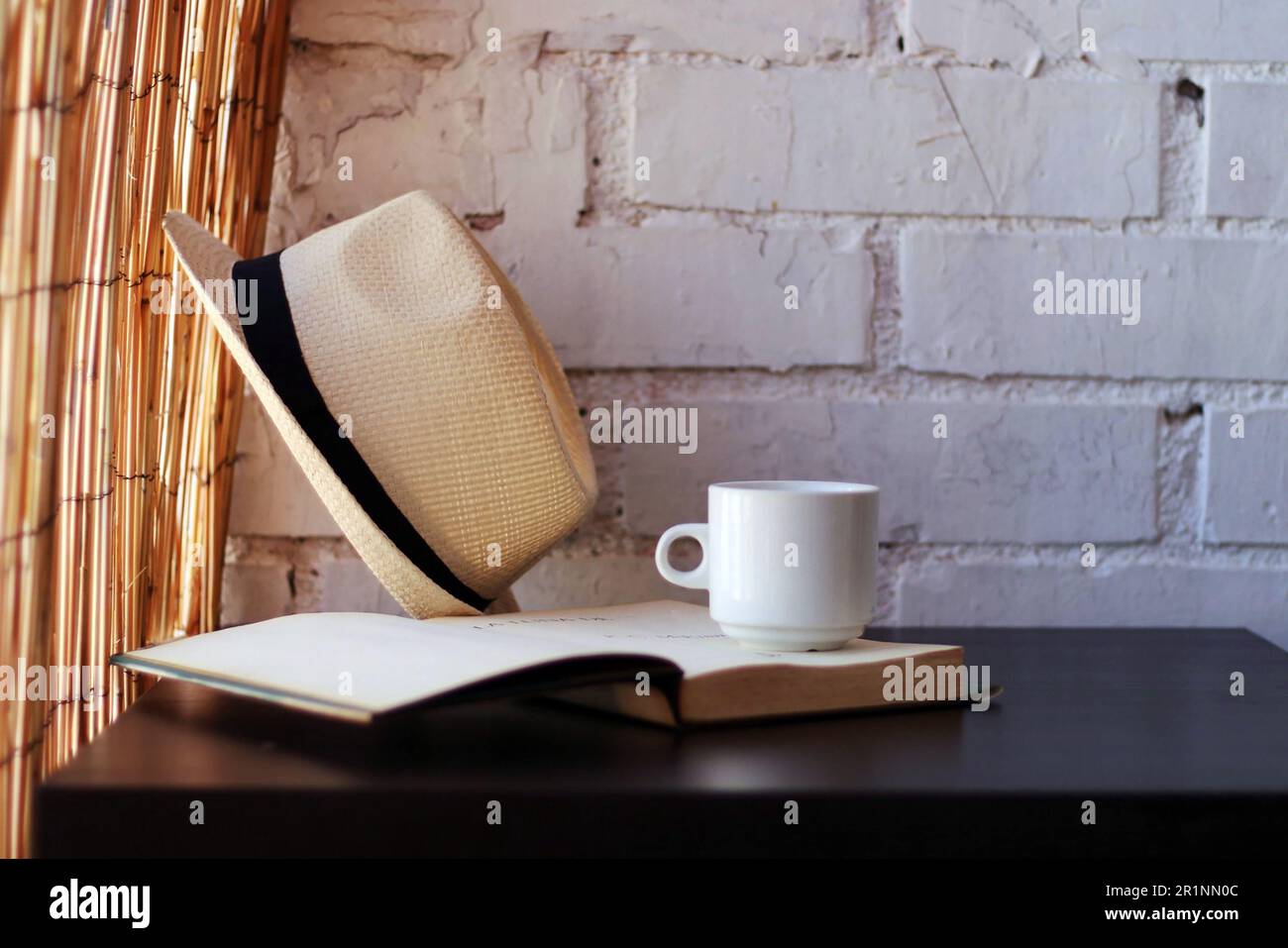 Becher, offenes Buch und Strohhut auf einem schwarzen Tisch. Stockfoto