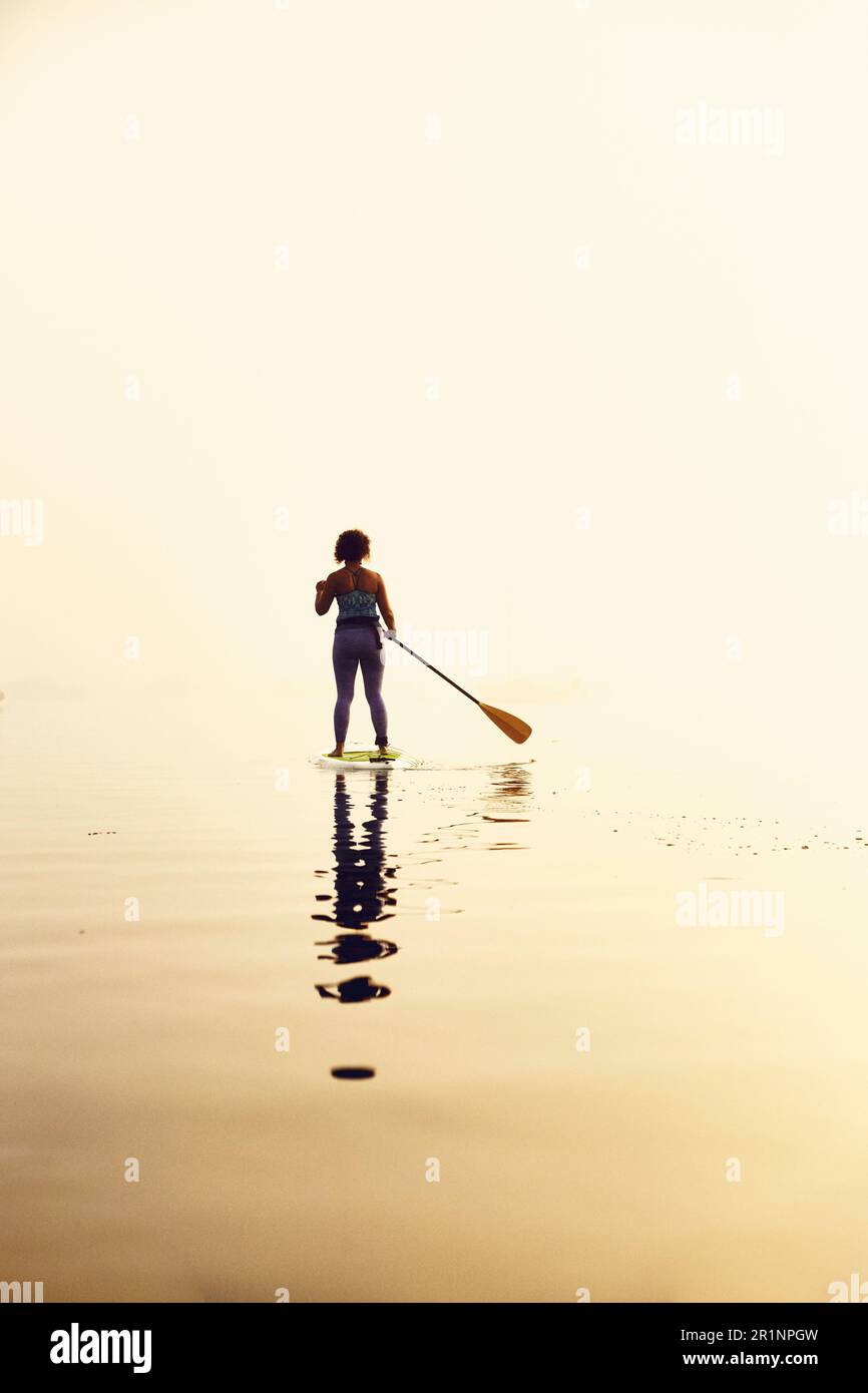 Junge Frau steht auf einem Stehpaddelboot an einem nebeligen Morgen bei Sonnenaufgang Stockfoto