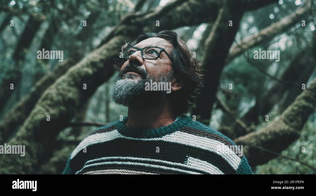 Porträt eines erwachsenen Mannes, der mit Waldbäumen im Hintergrund grüner Umgebung aufblickt. Nahaufnahme von männlichen Menschen in Naturpark Freizeitaktivitäten. Bart und Brille. Lebensumfeld Stockfoto