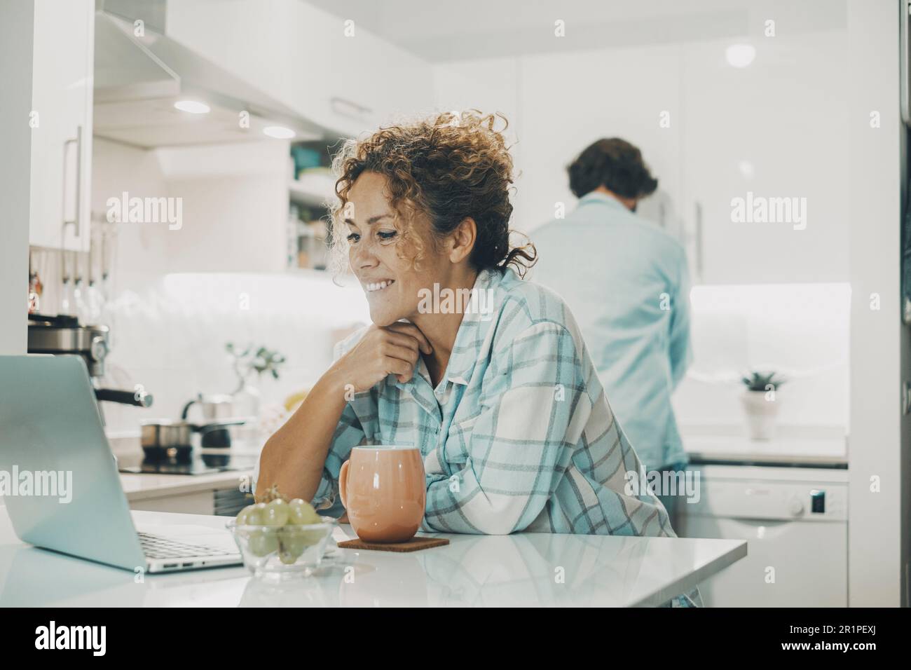 Ein glückliches Paar genießt die Zeit zusammen in der Küche zu Hause. Frau, ein Laptop, ein Mann, der im Hintergrund Geschirr spült. Beziehung und lebender Mann und Frau in der Wohnung Stockfoto