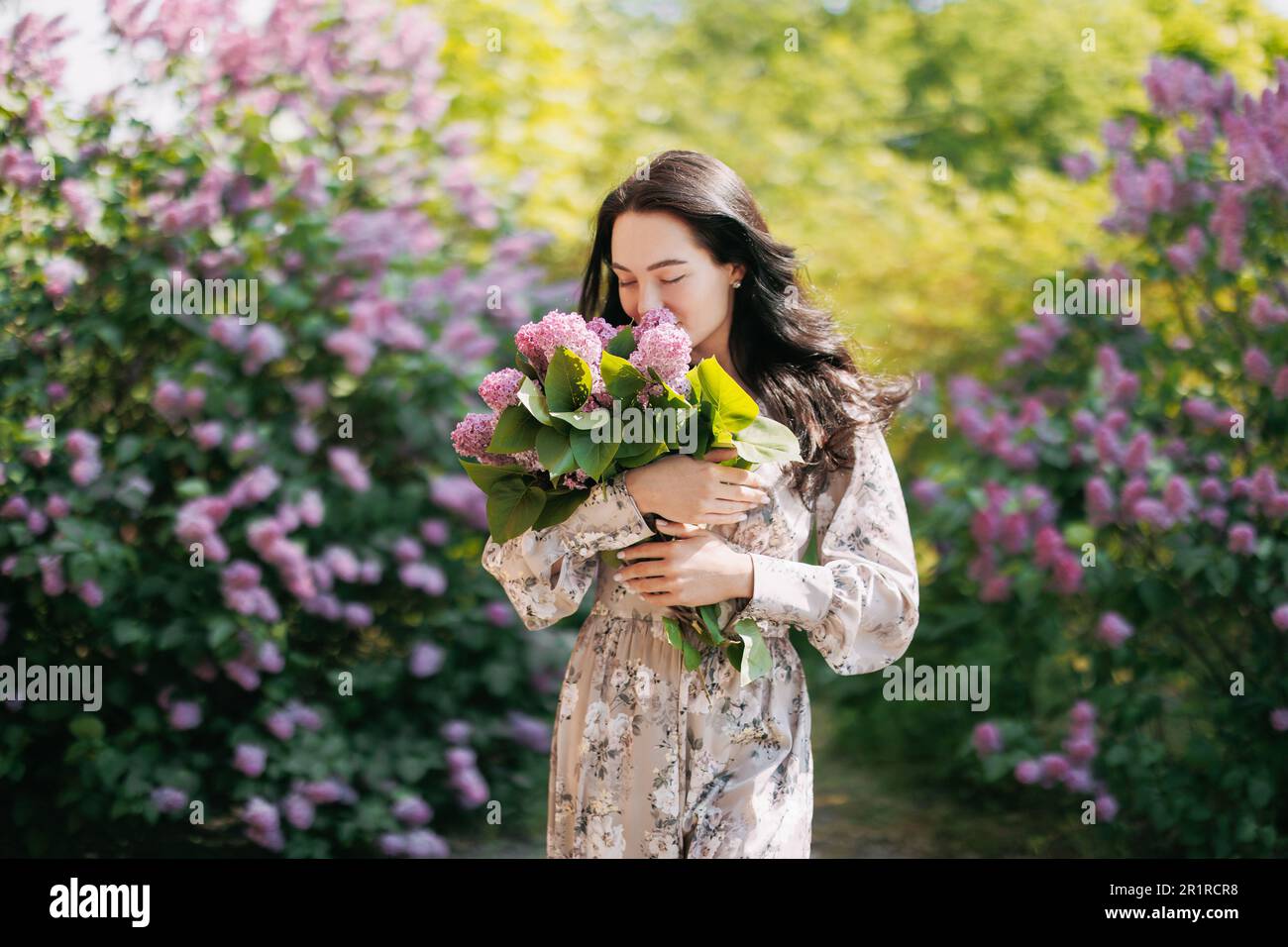 Die junge, hübsche, brünette Frau geht im Garten unter blühenden fliederroten Sträuchern spazieren und genießt den Blumenstrauß in ihren Händen . Stockfoto